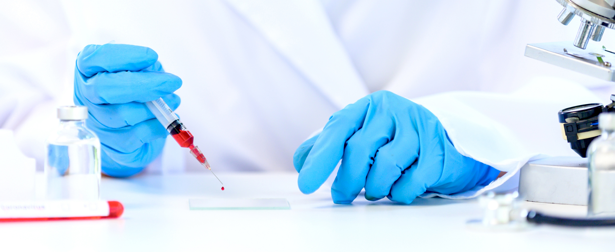 A laboratory technician wearing blue gloves uses a syringe to place a drop of blood onto a glass slide, with laboratory equipment visible on the table.