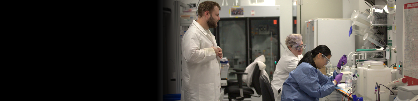 Scientist looks into a microscope with one scientist next to her and one behind her. 