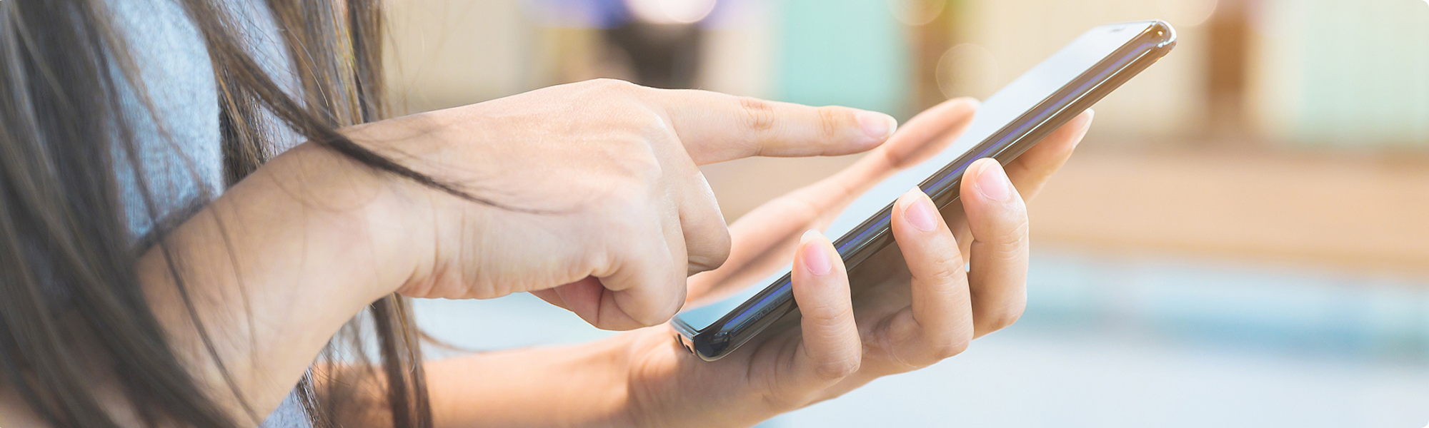 Photograph showing a person using a smartphone with one hand holding the device and the other hand tapping the screen. The background is blurred, emphasizing focus on the smartphone interaction.