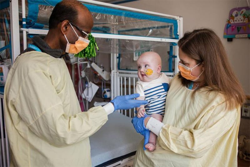 Two doctors, wearing masks, interact with a smiling infant patient. 