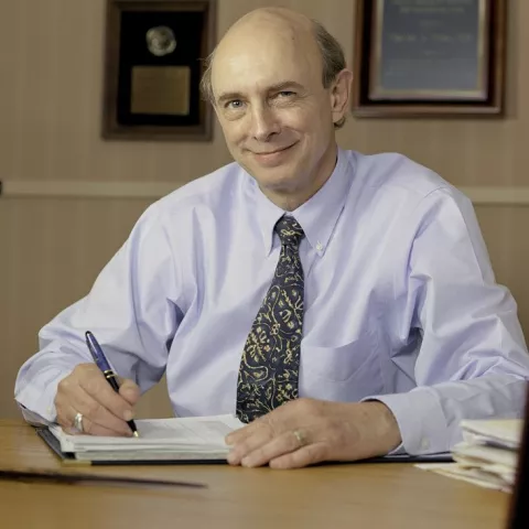 A man sits behind a desk while holding a pen and smiling at the camera.