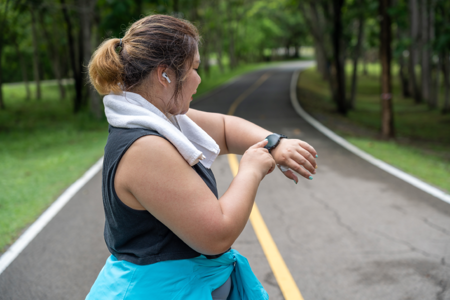 Young overweight woman checking the result of her morning run on her smart watch while standing at a running track of a local park.