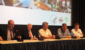 Five men seated at conference room with microphones, slide projected in background