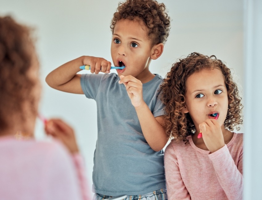 Two young children standing in front of a mirror, each brushing their teeth, showcasing a routine of oral care.