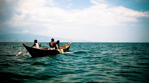 three young men in small wooden boat, fishing nets draped on side, wide expanse of blue water, mountains in distant background