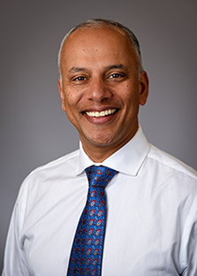 In this headshot of Dr. Chandy John he wears a white shirt and a predominantly blue paisley tie. 