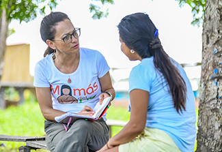 This photo shows Dr. Lisa Labita Woodson talking intently with a woman on a bench outdoors. Labita Woodson holds a pen and a notebook.