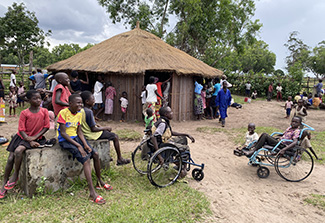Adults and children gather near the thatched-roofed health center (in the background) in the village of Kahemba. In the foreground, a group of children – many of them smiling – sit on a cement block, on the ground, or in wheelchairs.