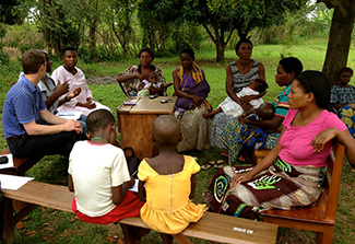 The photo shows a group of people, consisting of a couple of men and several women and children, sitting outside on wooden benches in a circle and engaging in discussion outside.