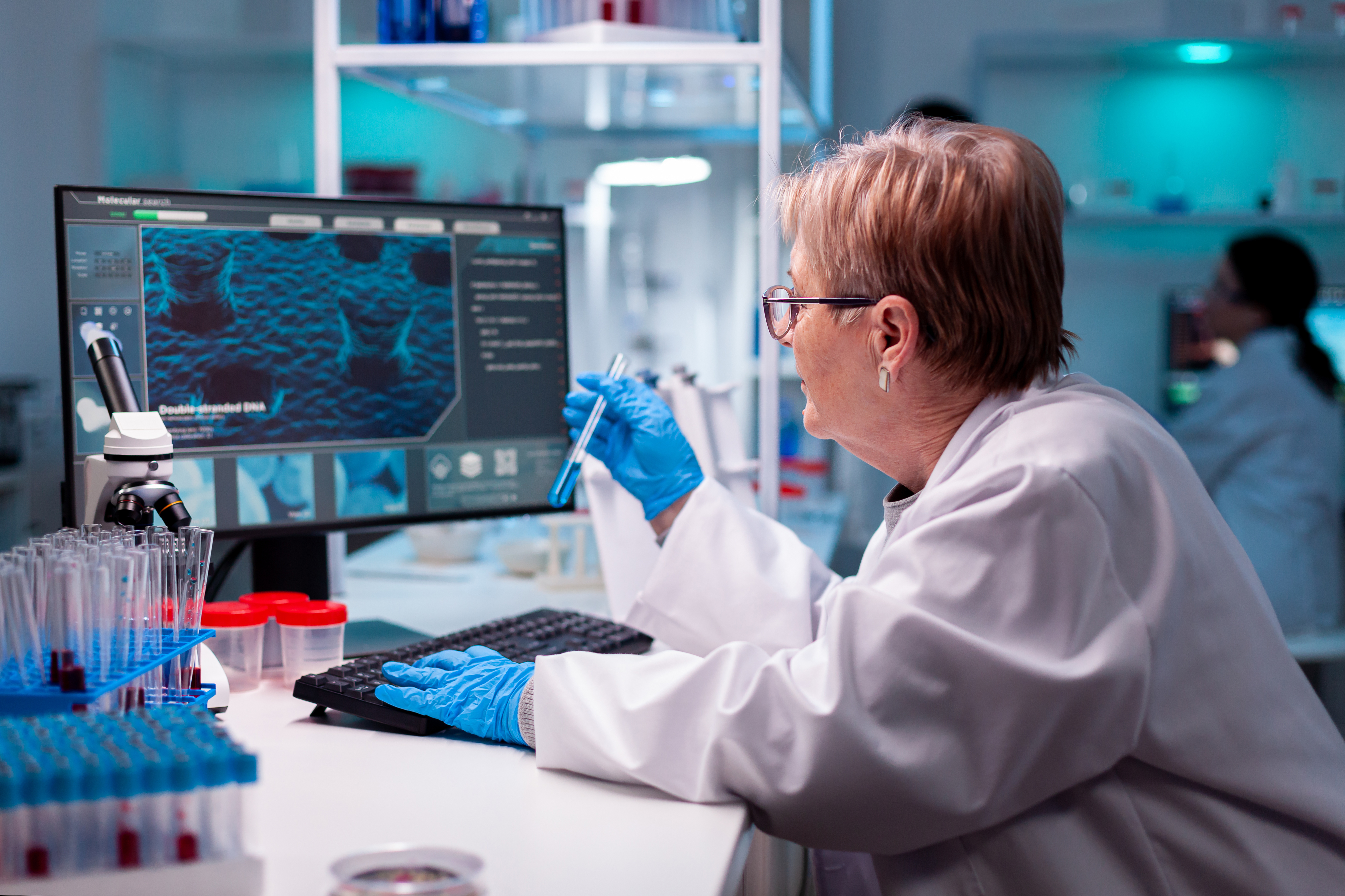 Scientist in lab coat holding test tube looks at computer screen