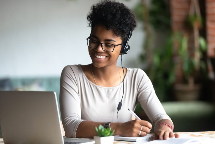 A person wearing headphones writes on paper while using a laptop.