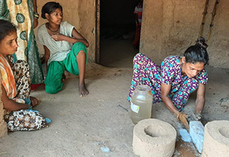 This photo shows two women, one is squatting behind next to a smokeless mud cookstove, the other is standing in front of the stove