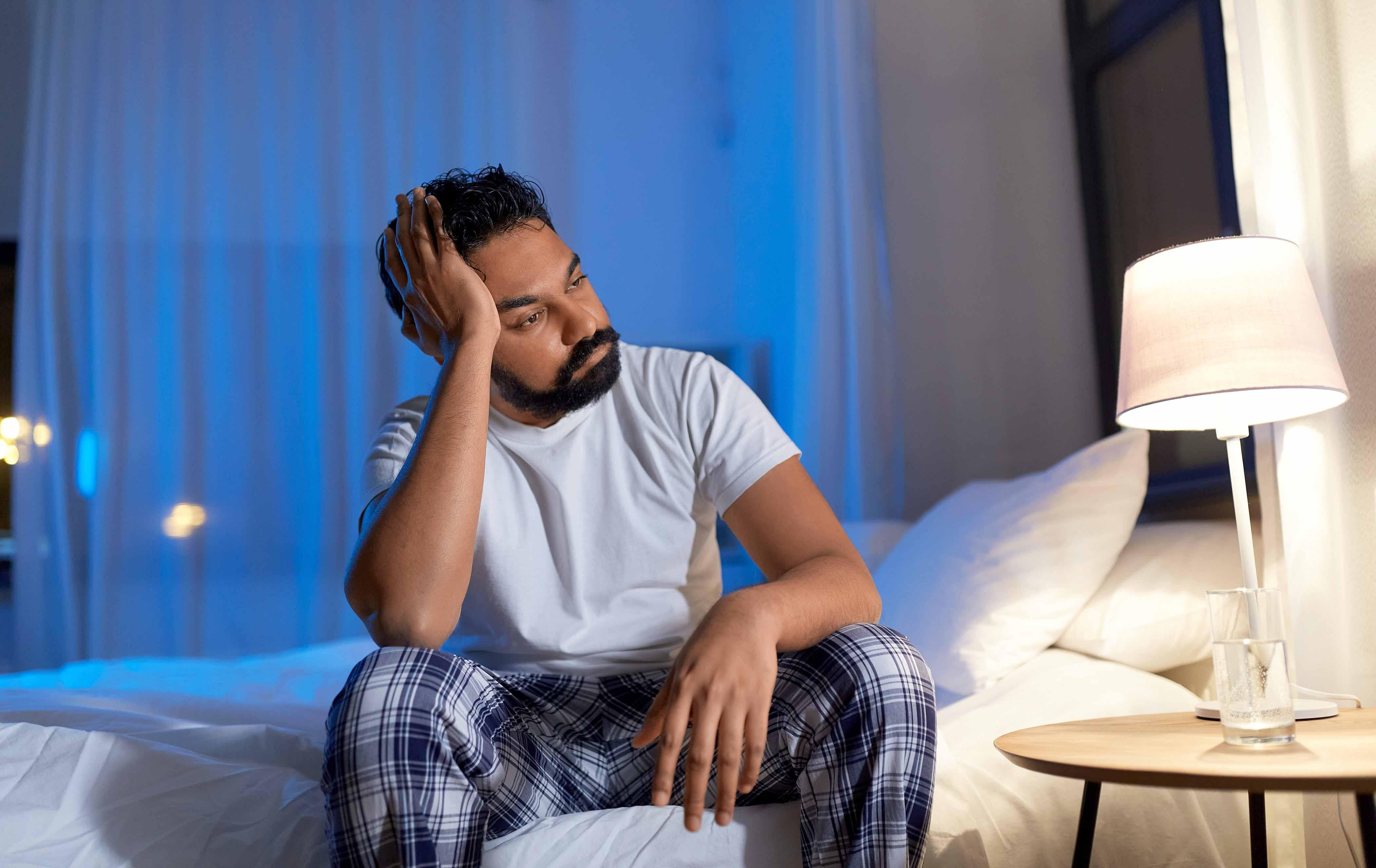 South Asian male sitting on the side of a bed, looking concerned.