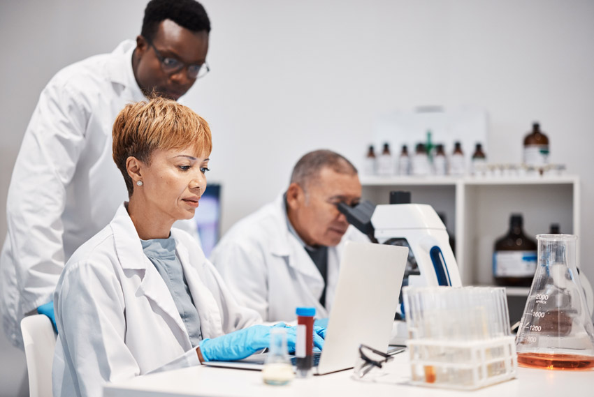 A female scientist sitting at a table and working on a computer next to a male scientist looking into microscope and a male scientist standing behind them