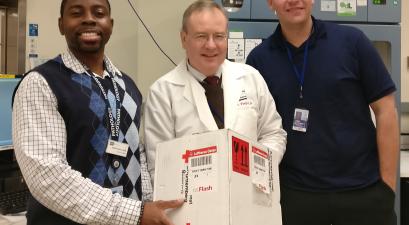Three smiling healthcare professionals stand together, with one holding a labeled medical package in a clinical or laboratory setting.