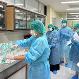A group of healthcare workers wearing teal surgical gowns, caps, and masks gather in a hospital or lab room, with one person handling supplies at a counter while others hold clipboards and papers, some taking notes, in front of glass-fronted cabinets stocked with medical supplies.